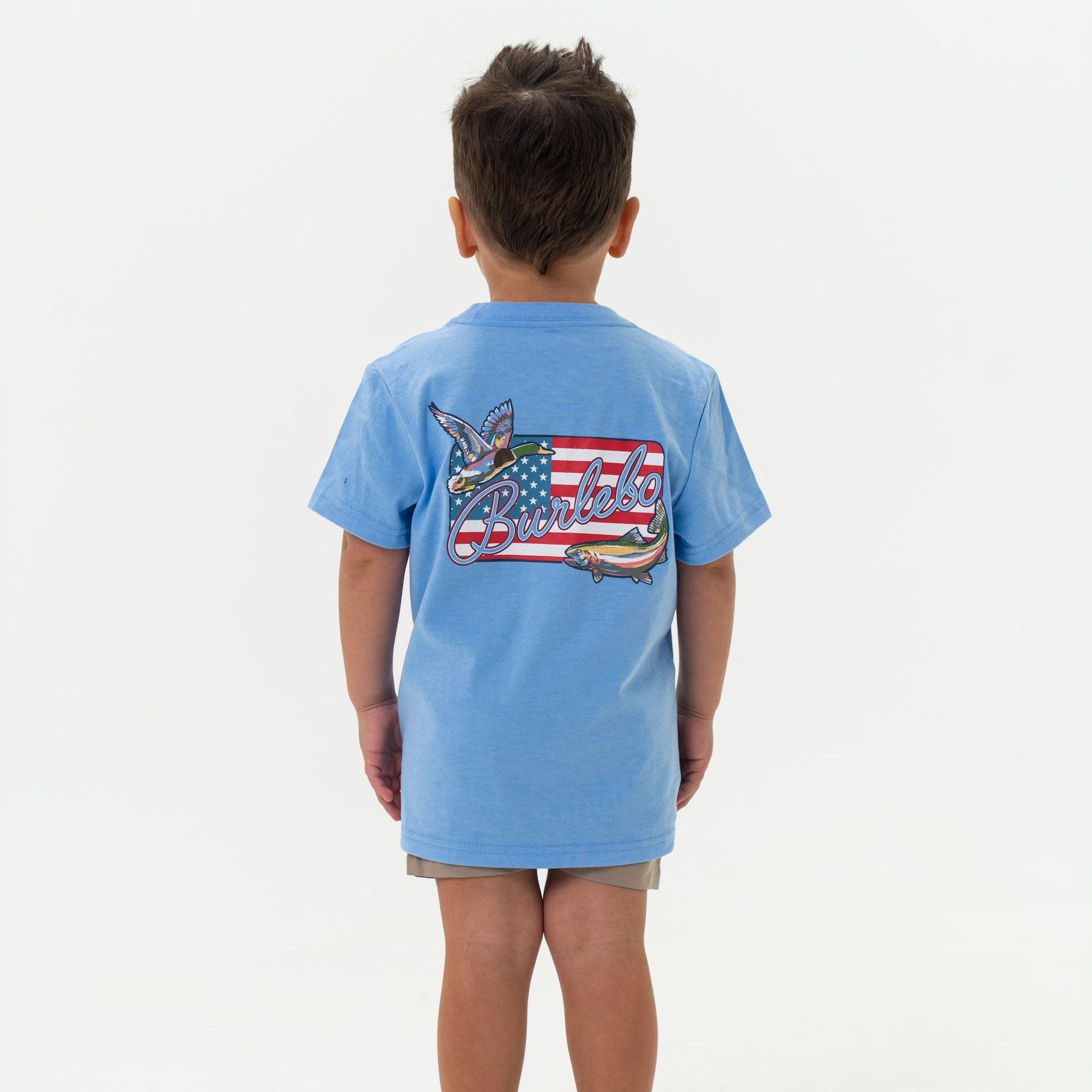 Burlebo Patch USA Youth Short Sleeve T-shirt, color light blue, youth sizes S-L, pre-shrunk, 50% cotton, 50% polyester.A young boy is shown from the back wearing a light blue short-sleeve T-shirt. The shirt features a colorful graphic on the back with an American flag background, mallard duck in flight, a fish, and the word "Burlebo" written in a bold, cursive font across the center. The boy is also wearing light khaki shorts and is standing against a plain white background.
